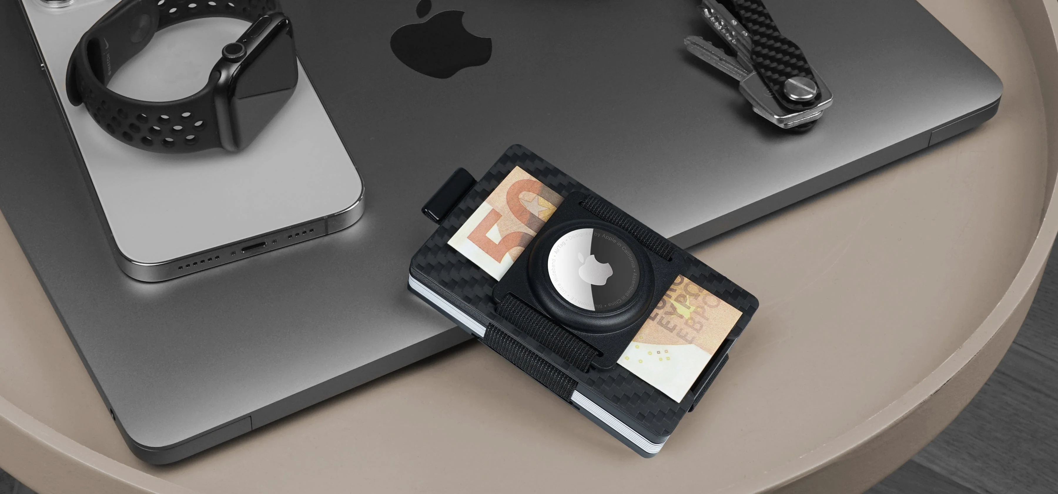 AirTag wallet on a desk with phone, watch, keys, and laptop for everyday carry